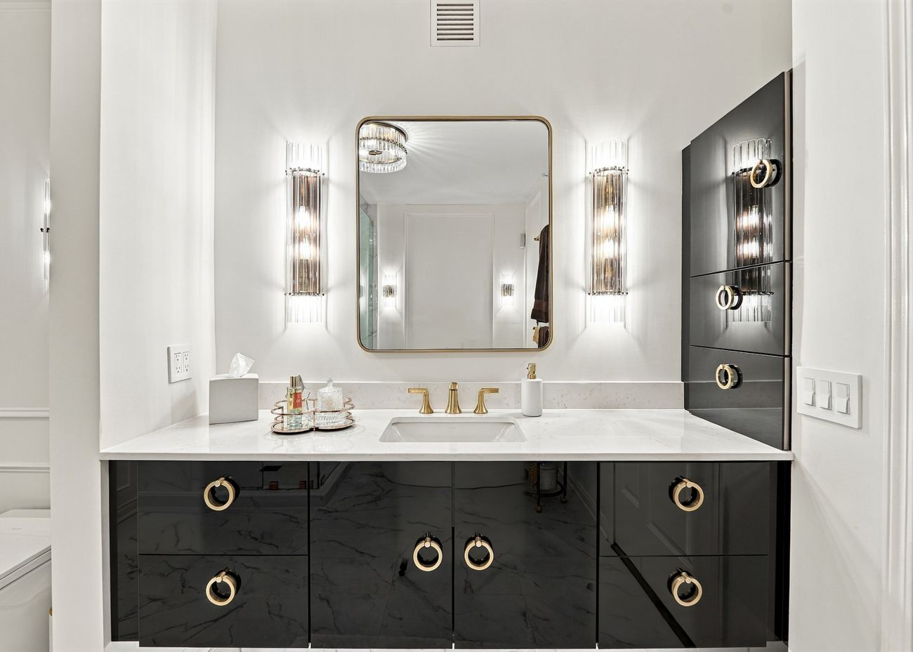 The same vanity after renovation: high-gloss black cabinetry with brass ring pulls, stone top and fluted sconces