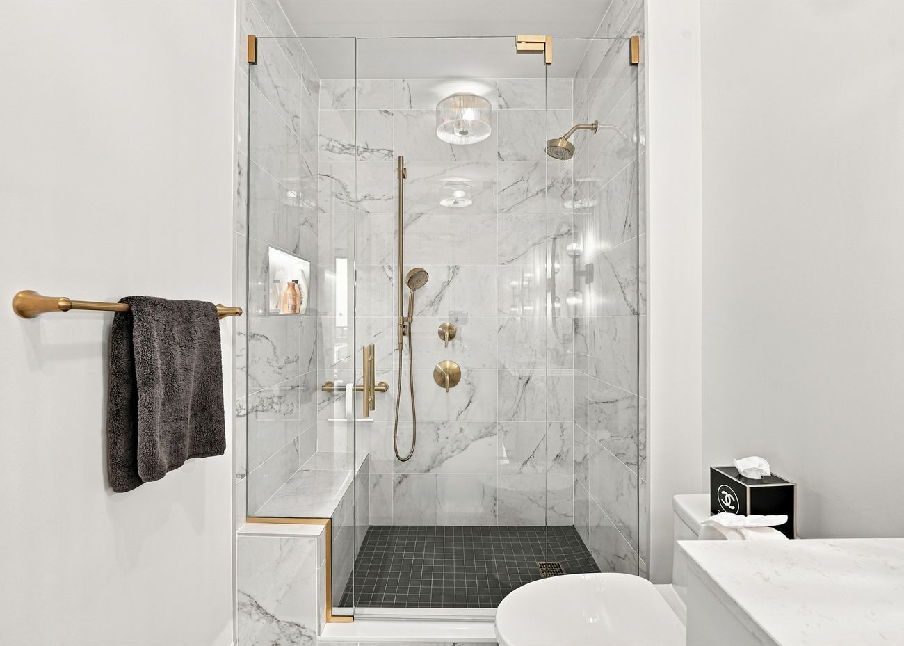 The same bathroom after renovation: marble walk-in shower with a dark hex floor and brass fittings